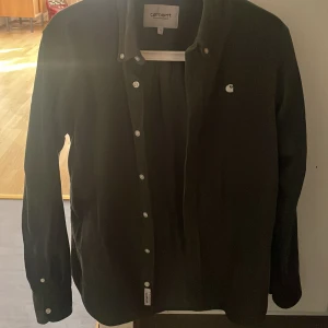 Mörkgrön  manchesterskjorta Carhartt S - Säljer en grå manchesterskjorta från Carhartt i storlek S. Skjortan har klassisk krage, knäppning framtill och vit broderad Carhartt-logga på bröstet. Långärmad och i mjuk manchester, perfekt för en chill och stilren look.