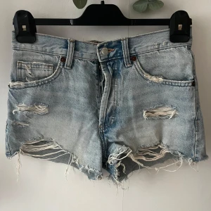 Ljusa slitna jeansshorts från Zara - Säljer ett par ljusblå jeansshorts från Zara med råa kanter och rejält slitna detaljer framtill och baktill. Klassisk femficksmodell med hög midja och avslappnad passform. Perfekta för en chill sommarlook.