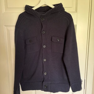 Mörkblå stickad kofta från pull and bear - Mysig mörkblå stickad kofta med huva och stora knappar framtill. Två bröstfickor med lock och ribbade muddar vid ärmslut och nederkant. Perfekt för kyliga dagar och enkel att slänga på över en t-shirt. Storlek L men passar perfekt till M