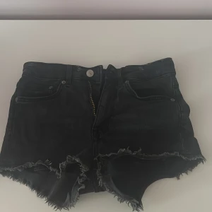 Svarta jeansshorts från H&M Divided - Svarta jeansshorts från H&M Divided i storlek 34. De har råa, fransiga kanter och klassisk femficksmodell. Shortsen har dragkedja och knapp framtill och är perfekta för en avslappnad och edgy stil.