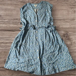 Blommig blå kortklänning med knappar - Supersöt blå kortklänning med små vita och gula blommor, knappar framtill och knytskärp i midjan. Ärmlös modell i mjukt bomullstyg, perfekt för varma dagar. Klänningen har en avslappnad passform och v-ringning.