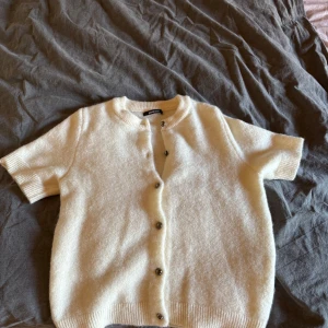 Vit kortärmad kofta från Gina Tricot - Superfin vit kofta från Gina Tricot med korta ärmar och rund hals. Mjuk stickad känsla och knappar framtill. Perfekt att slänga över ett linne eller klänning för en chill look. Ribbstickade muddar vid ärmslut och nederkant. Aldrig använd. Skriv om ni vill ha bilder när den är på 