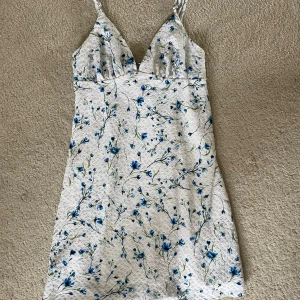 Vit blommig kortklänning med smala axelband - Superfin vit kortklänning med blått blommönster och smala axelband. Använd 2 ggr
