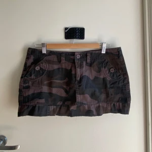 Camo minikjol med fickor - Snygg lowwaisted kortkjol i camouflage-mönster, man skulle kunna ta bort den lilla flared biten längs ner om man vill ha den ännu kortare. Den har två fickor med knappar på rumpan också.