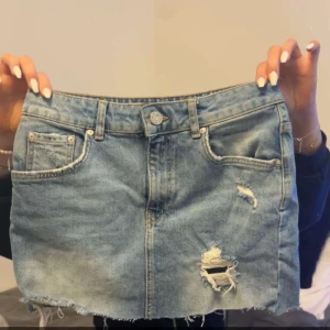 Blå jeanskjol med slitningar - Trendig blå jeanskjol med slitna detaljer och rå kant nertill. Klassisk femficksmodell med knapp och dragkedja framtill. Perfekt för dig som gillar en avslappnad och cool stil.
