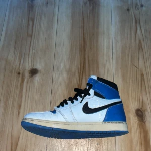 Nike Air Jordan 1 High Travis Fragment - Nike Air Jordan 1 High Travis Scott x Fragment med vit och blå ovandel i skinn, svarta detaljer och ikonisk omvänd swoosh. Blå yttersula och svarta skosnören. Perfekt för dig som vill sticka ut med en hypad sneaker.