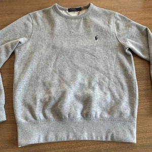 Grå sweatshirt från Polo Ralph Lauren - Klassisk grå sweatshirt från Polo Ralph Lauren med rund halsringning och broderad marinblå logga på bröstet. Tröjan har långa ärmar och ribbade muddar vid ärmslut och nederkant. Perfekt för en chill och stilren look. Säljer pga blev för liten 