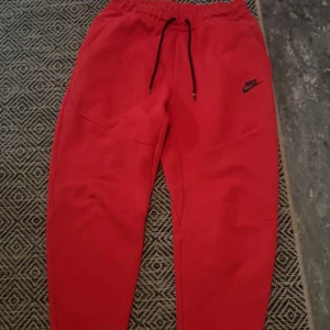 Röda mjukisbyxor från Nike - Röda mjukisbyxor från Nike med svart logga på benet och svarta snören i midjan. Byxorna har elastisk midja och är gjorda i mjukt bomullsmaterial, perfekta för chill eller träning. Snygg och enkel design som är lätt att matcha.