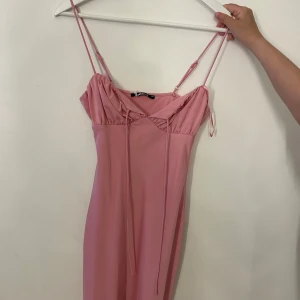 Rosa kortklänning från Gina Tricot - Supersöt rosa klänning från Gina Tricot med smala axelband och snygg v-ringning. Klänningen har en figurnära passform och fina knytband framtill. Perfekt för dig som gillar en feminin och trendig stil.