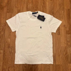 Vit t-shirt från Polo Ralph Lauren - Vit klassisk t-shirt från Polo Ralph Lauren med rund hals och korta ärmar. Ikonisk broderad logga i svart på bröstet. Tillverkad i mjuk bomull för en skön och stilren look. Perfekt till jeans eller shorts för en clean vibe.