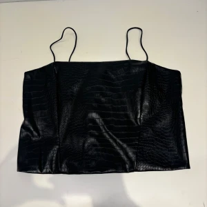 Svart croppad linne-topp i skinnimitation - Snygg svart croppad linne-topp med smala axelband och med krokodilpräglat mönster i skinnimitation. Toppen har en dragkedja baktill. 