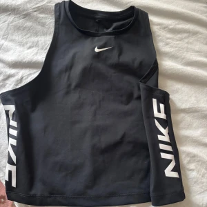 Svart Nike Pro Dri-FIT linnetopp - Sportig svart linnetopp från Nike Pro med Dri-FIT-teknologi. Toppen har en croppad passform, ärmlös design och stora vita Nike-loggor längs sidorna. Perfekt för träning och gym, tillverkad i ett stretchigt syntetmaterial som andas.