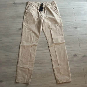Beige chinos från Tommy Hilfiger - Snygga beige chinos från Tommy Hilfiger med elastisk midja och dragsko med logga. Byxorna har raka ben, sidofickor och en bakficka. Tillverkade i mjuk bomull för en skön känsla och clean look. Perfekta för en chill och stilren outfit.