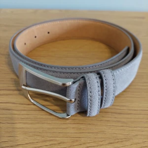 Grått mockabälte med silverfärgat spänne - Snyggt grått bälte i mocka med ett stilrent silverfärgat metallspänne. Bältet har en klassisk design och passar perfekt till jeans eller chinos. Enkel och modern look som lyfter din outfit.