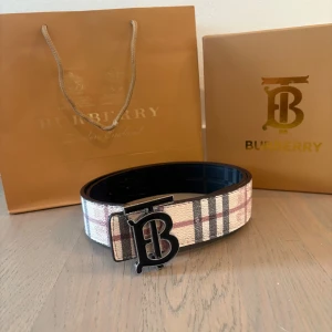 Burberry rutig bälte med B-spänne - Snyggt Burberry-bälte med klassiskt rutigt mönster i beige, svart och rött. Bältet har ett stort svart B-spänne framtill som ger en lyxig känsla. Perfekt accessoar för att lyfta din outfit med en ikonisk touch från Burberry.