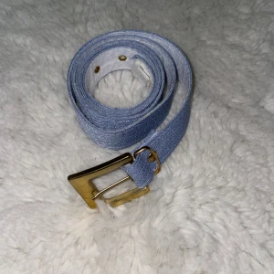Ljusblått jeansbälte med guldigt spänne - Snyggt ljusblått bälte i material som immiterar jeans och ett metallspänne i guldton. Bältet har flera hål för justerbar passform och en stilren, enkel design som passar till många outfits. Ca 105 cm långt. Liten fläck på insidan, se bild 4.