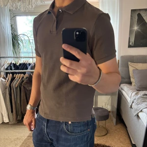 Brun pikétröja från H&M slim fit - Säljer en brun pikétröja från H&M i slim fit-modell. Tröjan har klassisk krage, korta ärmar och knäppning med två knappar. Materialet är mjukt och lätt strukturerat, perfekt för en clean och stilren look.