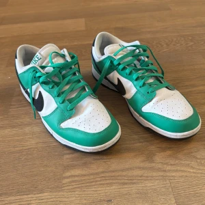 Nike Dunk Low vit/grön med svart swoosh - Nike Dunk Low sneakers i vitt och grönt med svart swoosh. Klassisk siluett med lågt skaft, gröna snören och perforerad tå. Ovandel i skinn och textil, svart yttersula och vit mellansula. Perfekt för dig som gillar streetwear och vill sticka ut.