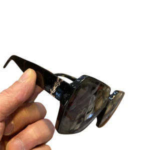 Svarta  solglasögon - Snygga svarta solglasögon med geometrisk, oversized form. Bågarna är i blank plast med guldigt logo på sidorna. Perfekt accessoar för en cool och trendig look.