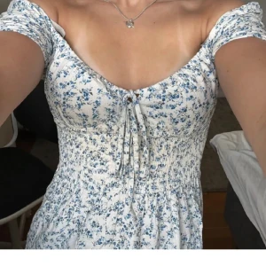 Vit blommig kortklänning med snörning - Supersöt vit kortklänning med blått blommönster och smockad midja. Klänningen har offshoulder-ärmar, v-ringning och snörning framtill. Perfekt för varma dagar och ger en romantisk vibe.