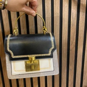 Svart och vit handväska med gulddetaljer - Snygg handväska i svart och vitt med guldfärgat handtag och lås. Väskan har dekorativa sömmar och en boxig form. Materialmix av skinn och textil, med coola detaljer som ger en lyxig vibe. Perfekt accessoar för att lyfta din outfit.