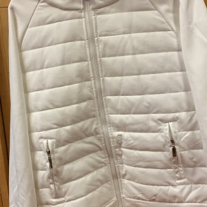 Ny vit quiltad pufferjacka med huva - Så fin vår/höst jacka, vit pufferjacka med horisontella quiltade paneler och dragkedja framtill. Jackan har två fickor med dragkedja och en skön huva. Perfekt för dig som gillar en clean och stilren look. Passar till både jeans och mjukisbyxor. Helt oanvänt pga av fel storlek. Det är en liten L. 
