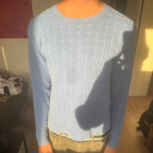 Ljusblå kabelstickad tröja - Säljer en ljusblå kabelstickad tröja med rund hals och långa ärmar. Tröjan har klassiskt stickat mönster och ribbade muddar vid ärmslut och nederkant. Perfekt för dig som gillar en clean och stilren look.