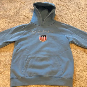 Blå hoodie från GANT med logga - Snygg ljusblå hoodie från GANT med broderad logga och sköld på bröstet. Klassisk känguruficka framtill och huva. Perfekt för dig som gillar stilren och sportig look. Tillverkad i mjuk bomull för extra komfort.