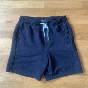 Mörkblå shorts med snörning - Säljer ett par mörkblå shorts med resår i midjan och vit snörning framtill. Shortsen har en bakficka och är tillverkade i ett mjukt, strukturerat material. Perfekta för sommardagar eller avslappnade tillfällen.