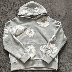 Grå hoodie från Denim Tears med blommor - Säljer en grå hoodie från Denim Tears med vita blommiga tryck över hela tröjan. Tröjan har huva, magficka och ribbade muddar. Perfekt för dig som gillar streetwear och vill sticka ut med en unik design. Har bara använt den en gång och känner att detta inte riktigt är min stil så vill sälja den 