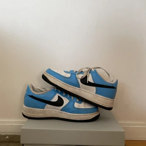 Nike Air Force 1 blå och vita sneakers - Nike Air Force 1 sneakers i blått och vitt med svarta detaljer och klassisk svart swoosh. Låga med snörning, perforerad tå och vit mellansula. 