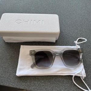  Solglasögon från CHIMI - Snygga solglasögon från CHIMI med transparent grå båge och fyrkantig form. Glasen är mörktonade och bågarna har CHIMI-logga på sidorna. Kommer med originalfodral, putsduk och påse. Perfekta för dig som vill ha en clean och modern look.