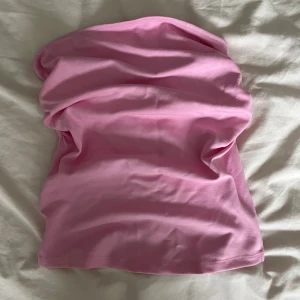 Rosa bandeau tubtopp från Bershka.  - Säljer en ljusrosa bandeau tubtopp från Bershka. Toppen är ärmlös och har en enkel, slät design med en liten rynkdetalj framtill. Tillverkad i mjuk bomullsmix som sitter skönt mot kroppen. Perfekt för dig som gillar en clean och trendig look. Den har två små svarta fläckar.