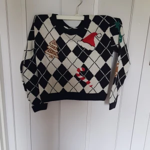 Svartvit jultröja med mönster från H&M - Snygg svartvit stickad tröja från H&M med rutigt mönster och juliga detaljer som tomteluva, pepparkaka, polkagris och julgranskulor. Långärmad och perfekt för vinterns mysiga dagar. Tröjan har en klassisk passform och är riktigt skön att ha på sig.