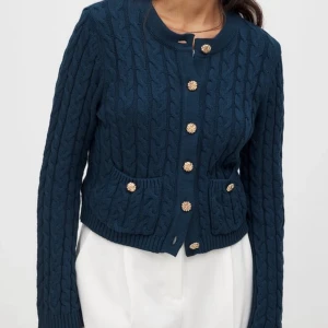 Mörkblå kabelstickad kofta med guldknappar - Säljer en mörkblå kabelstickad kofta med snygga guldiga knappar framtill och på fickorna. Koftan har rund halsringning, ribbade muddar och två fickor fram. Perfekt för lager-på-lager och ger en chic vibe till din outfit.