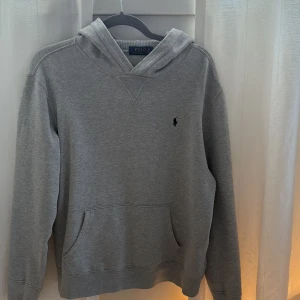 Grå hoodie från Polo Ralph Lauren - Klassisk grå hoodie från Polo Ralph Lauren med broderad logga på bröstet. Tröjan har huva, känguruficka och långa ärmar. Tillverkad i mjuk bomullsmix som är perfekt för chill dagar. Enkel och stilren design som funkar till allt.