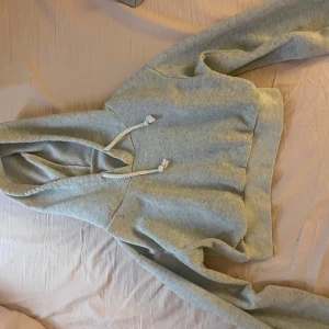 Ljusgrå croppad hoodie med huva - Säljer en ljusgrå croppad hoodie med huva och vita snören. Tröjan har långa ärmar och bred mudd nertill för en snygg passform. Perfekt för en chill och avslappnad stil.