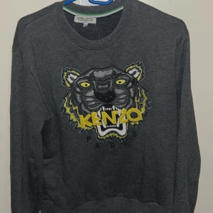 Kenzo tröja  - Säljer en grå sweatshirt från Kenzo med ikoniskt broderat tigerhuvud och gult Kenzo Paris-tryck på bröstet. Tröjan har rund halsringning och långa ärmar. Perfekt statement piece för dig som vill sticka ut. S men passar M
