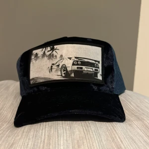 Blå keps med Lamborghini  - Snygg keps från Hatstore med mesh baktill. Framsidan har ett stort tygmärke med en svartvitt bild av en lamborghini och palmer som gör den väldigt snygg. Skärmen och fronten är i sammetsliknande material för extra stil. Passar bra till sommaren då man kan behöva en keps för skydd och varför inte ha en snygg keps isåfall. Svin bra skick. 