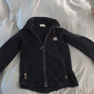 Svart fleecejacka från Moncler - Svart fleecejacka från Moncler med dragkedja framtill och två fickor med dragkedja. Jackan har hög krage och klassisk Moncler-logga på bröstet. Tillverkad i mjuk polyester som är perfekt för kyliga dagar. Enkel och stilren design som funkar till det mesta. Jag säljer denna för att den är för liten för mig. Jag har ungefär storlek 12-13 men den passar mig och den är storlek 10. Jag har använt den några gånger, men den är i väldigt bra skick. Det är äkta moncler, kontakta om ni har några frågor 💕 