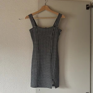 Svartvit rutig kort klänning - Snygg svartvit rutig kortklänning med breda axelband och liten slits framtill. Figurnära passform som ger en cool och trendig look. Perfekt för dig som gillar klassiska mönster med en modern twist.🥰