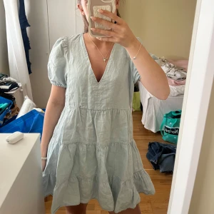 Ljusblå kort klänning i linne - Supersöt ljusblå klänning med v-ringning och puffärmar. Den har volanger i flera lager och är sydd i ett luftigt linnetyg, perfekt för varma dagar. Klänningen är kort och har en avslappnad passform som ger en chill vibe.