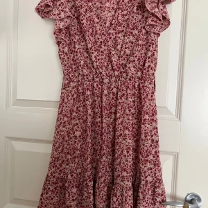 Blommig röd kortklänning med volang - Supersöt kortklänning med rödblommigt mönster och volangärmar. Klänningen har omlott-look upptill, markerad midja och volangdetaljer nertill. Perfekt för sommarens alla tillfällen. Lätt och luftig känsla.