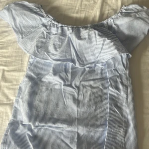 Blåvit randig blus med volang - Säljer en söt blus med blå och vita smala ränder och stor volang runt axlarna. Blusen har offshoulder-stil och är luftig med en lös passform, perfekt för varma dagar.