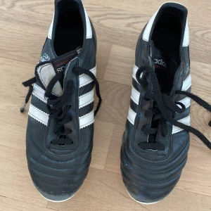 Adidas Copa Mundial fotbollsskor svart/vit - Klassiska Adidas Copa Mundial fotbollsskor i svart skinn med vita detaljer och de ikoniska tre ränderna på sidorna. Skorna har snörning och fasta dobbar för gräsplan. Perfekta för dig som vill ha en tidlös och stilren look på planen.