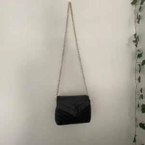 Svart quiltad axelväska med kedja - Säljer en svart quiltad axelväska i skinnimitation med snygg guldfärgad kedjerem. Väskan har lockstängning med magnetknapp och ett huvudfack med innerficka med dragkedja. Perfekt storlek för mobil, plånbok och småprylar.