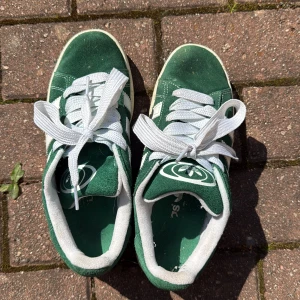 Adidas Campus gröna sneakers 40 - Gröna Adidas Campus sneakers i mocka med klassiska vita ränder och vit sula. Skorna har vit snörning, gummisula i ljusbrun färg och rund tå. Perfekta för dig som gillar retrostil och streetwear.