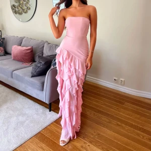 Ljusrosa långklänning med volanger - Superfin ljusrosa långklänning med axelbandslös design och massor av volanger längs kjolen. Klänningen har en snygg slits framtill och sitter tight upptill för en elegant look. Perfekt för dig som vill sticka ut med en romantisk vibe. Har endast använt den en gång, ordinarie pris 1400