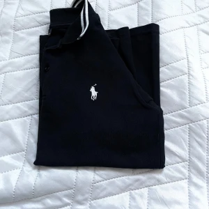 Svart pikétröja från Ralph Lauren - Klassisk svart pikétröja från Ralph Lauren med vit broderad logga på bröstet. Tröjan har korta ärmar, krage med vita ränder och knäppning framtill. Perfekt för en clean och sportig stil. Tillverkad i mjuk bomull som andas. Passar personer som är ca 150-160cm långa