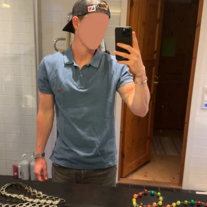 Blå pikétröja - Säljer en klassisk blå pikétröja med korta ärmar och krage. Tröjan har en liten röd logga broderad på bröstet och är gjord i mjuk bomull. Perfekt för en avslappnad och stilren look.